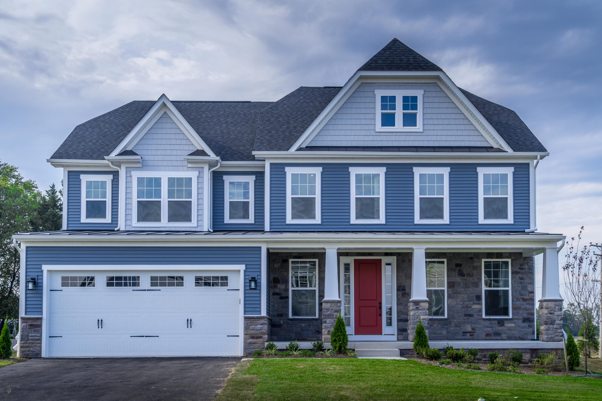 Two-story house with a garage, blue siding, red door, and landscaped yard under a cloudy sky